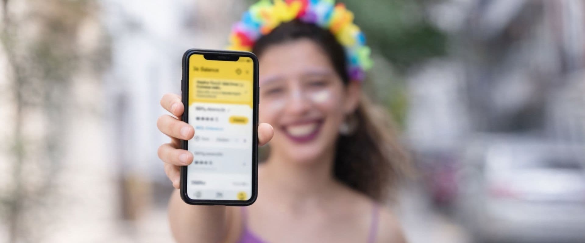Jovem sorridente usando um celular com uma tela amarela, em fundo urbano, celebrando o sucesso de um aplicativo ou serviço digital.